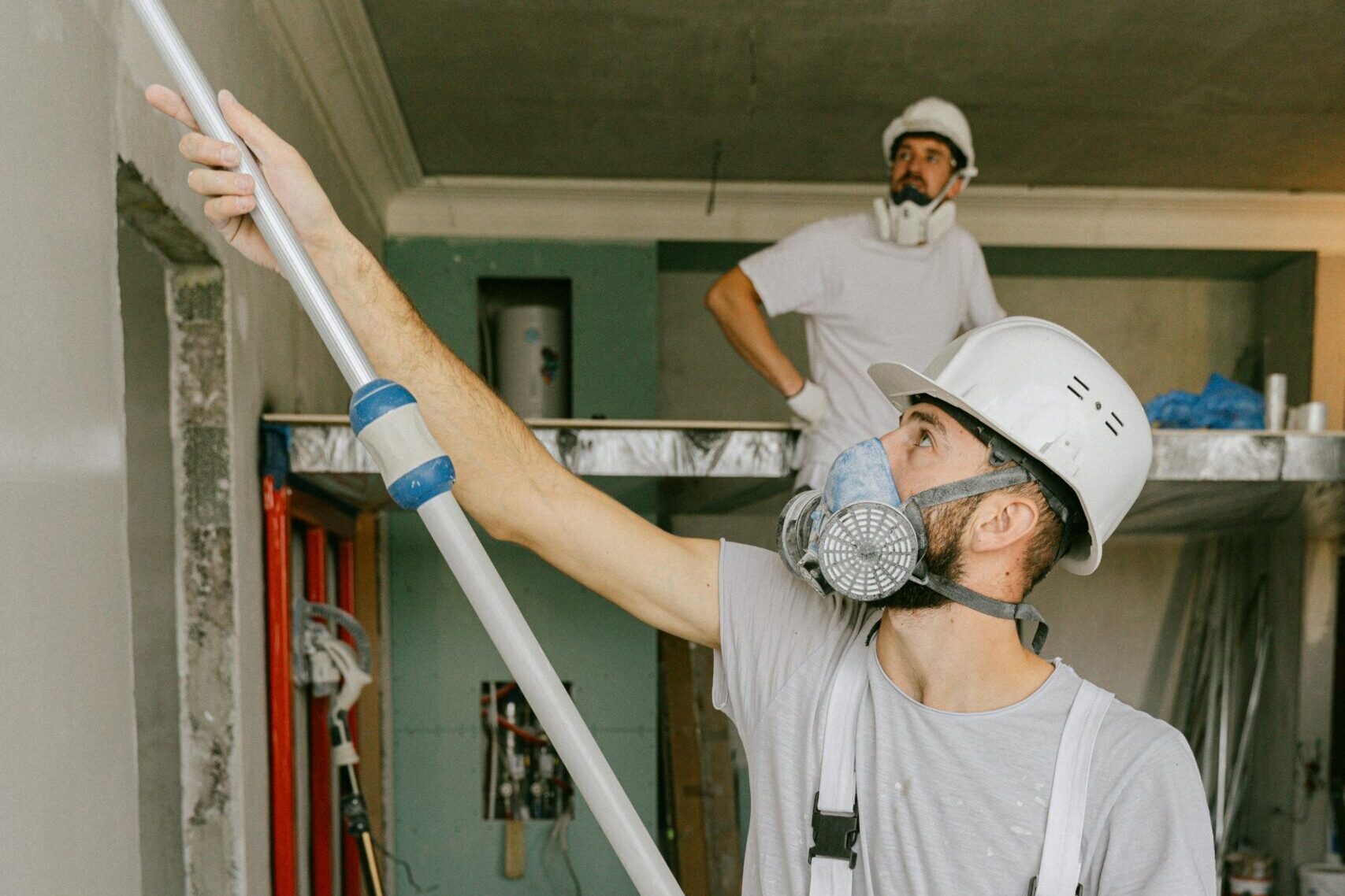 Two construction workers in safety gear are interior renovating, using a paint roller.